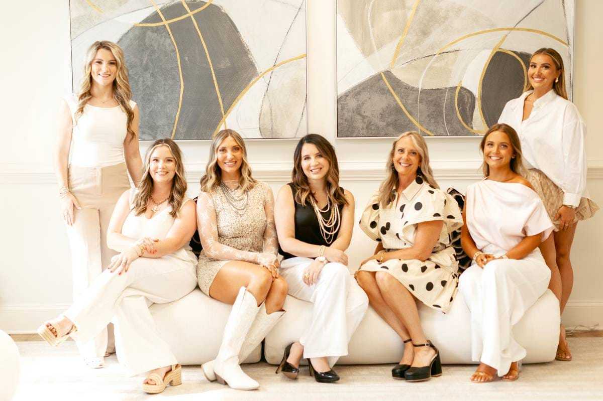 Seven women smiling in stylish outfits, seated in a bright room with abstract art.